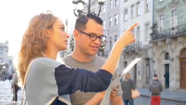 Ευτυχισμένοι νέοι τουρίστες ζευγαράκι που κατέχουν ένα έγγραφο χάρτη της αρχαίας ευρωπαϊκής πόλης νωρίς το πρωί σε κενή πλατεία και δείχνοντας σε κάποια αξιοθέατα μέρος — Αρχείο Βίντεο