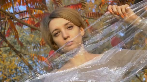 Junge Frau mit kurzen blonden Haaren blickt durch eine Plastiktüte, Polyethylenfolie, die draußen an einem sonnigen Herbsttag in der Nähe des Baumes mit gelben und roten Blättern steht. — Stockvideo