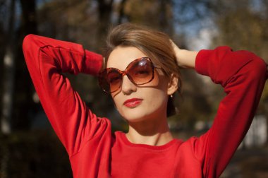 Güneşli bir günde sokakta poz veren çekici, güneş gözlüklü, çekici, kısa saçlı, şehvetli dudaklı ve kırmızı elbiseli genç kadın portresi.
