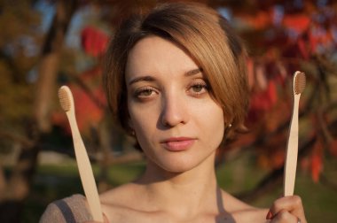 Kısa sarı saçlı genç kız, sonbaharda kırmızı ve turuncu arka planda güneşli havada dışarıda bambu diş fırçası tutuyor. Çevresel dostluk ve sıfır atık kavramı