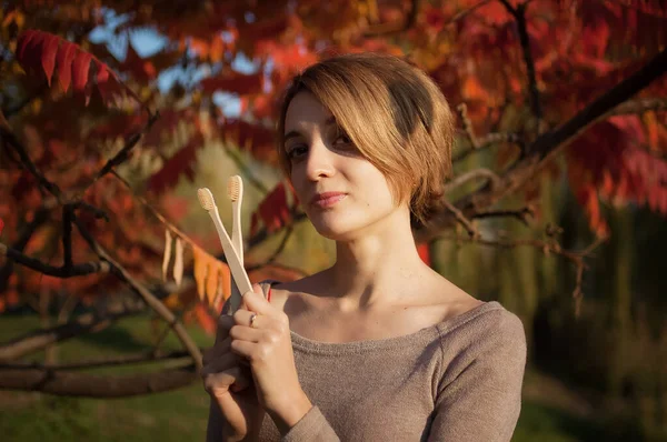 Ung flicka med kort blont hår håller en bambu tandborstar utomhus under soligt väder på hösten på röda och orange löv bakgrund. Miljövänlighet och nollavfallsbegreppet — Stockfoto