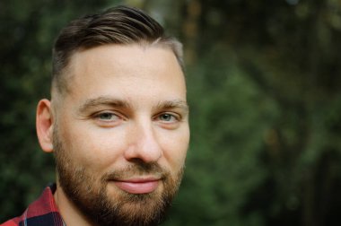 Kırmızı tişörtlü genç sakallı adamın portresini yakın çekimde ağaçların arkasındaki parktaki gülümsemeyle kameraya bakarak.