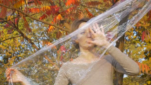 Mujer joven con el pelo corto y rubio está mirando a través de una bolsa de plástico, envoltura de polietileno de pie al aire libre durind día soleado otoño cerca del árbol con hojas amarillas y rojas . — Vídeos de Stock