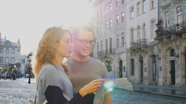 Feliz casal de jovens turistas segurando um mapa de papel da antiga cidade europeia no início da manhã na praça vazia e apontando para algum lugar de turismo — Vídeo de Stock