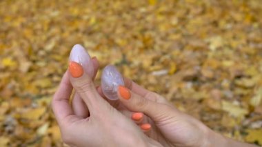 Turuncu manikürlü bir kadın elinde pembe kuvarstan yapılmış iki yoni yumurtası ve sonbaharda düşen sarı yapraklar üzerinde vumfit, imbuilding veya meditasyon için saydam menekşe ametist taşıyor.