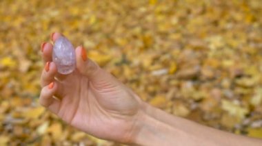 Turuncu manikürlü kadın eli, sonbahar günü açık havada düşen sarı yapraklar üzerinde vumfit, imbuilding veya meditasyon için şeffaf menekşe ametist yoni yumurtası tutuyor.