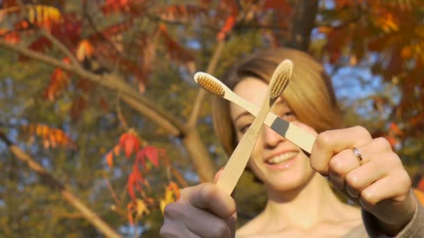 Jong meisje met kort blond haar houdt een bamboe tandenborstels buiten bij zonnig weer in de herfst op rode en oranje bladeren achtergrond. Milieuvriendelijkheid en nulafvalconcept — Stockvideo