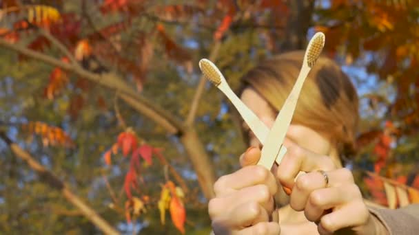 Chica joven con el pelo corto y rubio está sosteniendo un cepillo de dientes de bambú al aire libre durante el clima soleado en otoño sobre fondo de hojas rojas y naranjas. Adecuación al medio ambiente y concepto cero residuos — Vídeo de stock