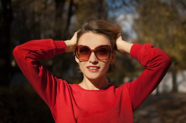 Güneş gözlüğü takan ve kırmızı kazak giyen güzel bir kadın açık havada güneşli bir havada sonbahar parkında poz veriyor.