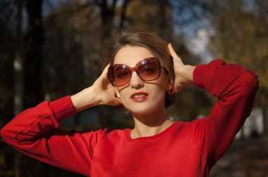 Güneş gözlüğü takan ve kırmızı kazak giyen güzel bir kadın açık havada güneşli bir havada sonbahar parkında poz veriyor.