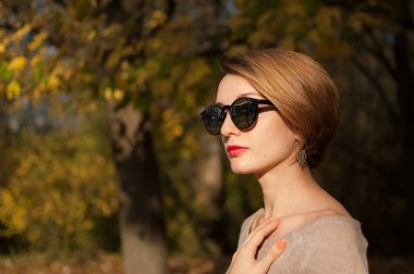 Kısa saçlı, güneş gözlüklü, sarı yapraklı ağaçlara poz veren genç ve güzel bir kız sonbahar parkında vakit geçiriyor.