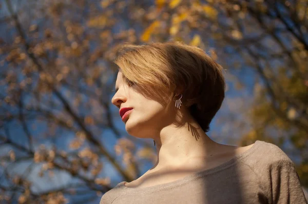 Draußen Porträt einer schönen jungen Dame mit kurzen Haaren, die silberne Ohrringe und Biege-Kleid trägt und vor blauem Himmel und den Bäumen mit gelben Blättern posiert, die Zeit im herbstlichen Park verbringt — Stockfoto