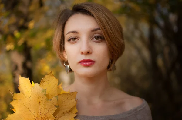 Atractiva joven con el pelo corto rubio y pendientes de plata sosteniendo un ramo de hojas de otoño en un hermoso parque de oro rojo al aire libre — Foto de Stock