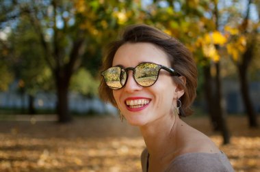 Kısa saçlı, güneş gözlüklü, sarı yapraklı ağaçlara poz veren genç ve güzel bir kız sonbahar parkında vakit geçiriyor.