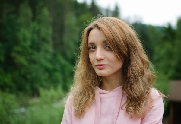 Atractiva joven mujer rubia tranquila en ropa rosa con pelo largo en la colina con fondo de bosque verde durante la primavera o principios de otoño en las montañas . — Foto de Stock