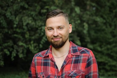 Kırmızı tişörtlü genç sakallı adamın portresini yakın çekimde ağaçların arkasındaki parktaki gülümsemeyle kameraya bakarak.