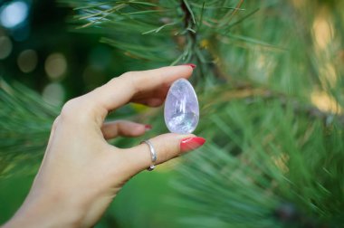 Vumfit, imbuilding veya meditasyon için şeffaf ametist kuvars yoni yumurta ile Kadın el. Çam ağacı dalları arka plan açık havada ellerde parlayan kristal yumurta. Kadın sağlığı kavramı