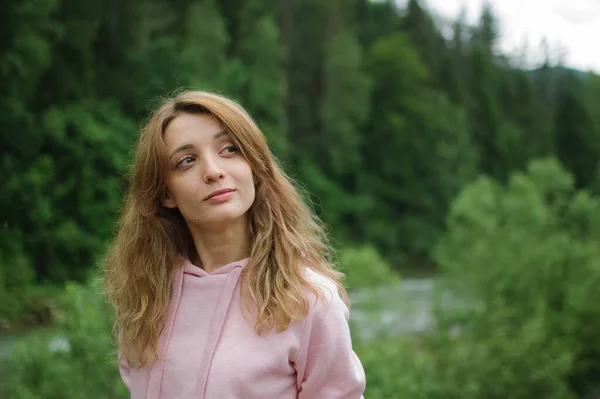 Increíble chica joven en ropa casual rosa con pelo largo en la colina con fondo de bosque verde durante la primavera o principios de otoño en las montañas . — Foto de Stock