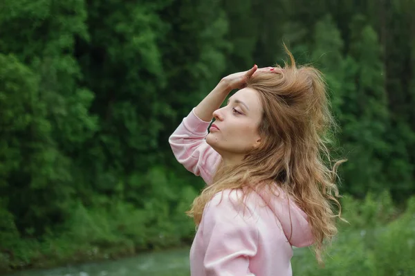 Vue latérale de la jeune fille étonnante en vêtements décontractés roses avec les cheveux longs et le sourire charmant sur la colline avec fond de forêt verte au printemps ou au début de l'automne dans les montagnes . — Photo