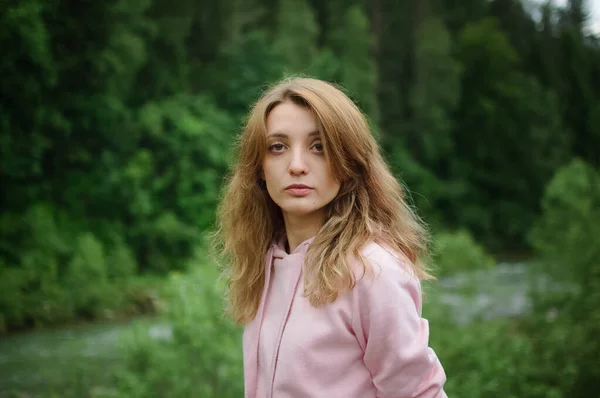 Mulher loira jovem grave em roupas rosa com cabelos longos está olhando para a câmera com ansiedade na colina com fundo verde floresta durante a primavera ou início do outono nas montanhas . — Fotografia de Stock