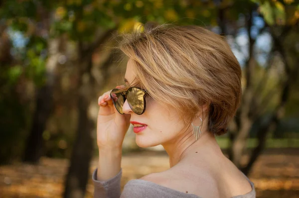 Młoda i piękna dziewczyna z krótkimi włosami i okularami przeciwsłonecznymi z lustrzaną powierzchnią pozuje na drzewa z żółtymi liśćmi tła spędzając czas w jesiennym parku — Zdjęcie stockowe