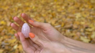 Turuncu manikürlü kadın eli, sonbahar günü açık havada düşen sarı yapraklar üzerinde vumfit, imbuilding veya meditasyon için pembe kuvars yumurtası tutuyor.