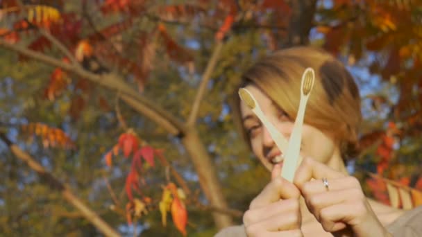 Kısa sarı saçlı genç kız, sonbaharda kırmızı ve turuncu arka planda güneşli havada dışarıda bambu diş fırçası tutuyor. Çevresel dostluk ve sıfır atık kavramı — Stok video