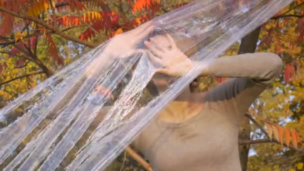 Mladá žena s krátkými blond vlasy se dívá přes plastový sáček, polyethylen zábal stojí venku durind slunečný podzimní den v blízkosti stromu se žlutými a červenými listy. — Stock video