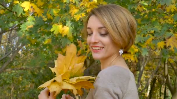 Емоційно приваблива молода жінка з коротким волоссям і сукнею, що тримає букет осіннього листя над головою і позує в красивому парку на відкритому повітрі — стокове відео