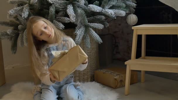 Nettes blondes Mädchen hat eine Geschenkschachtel in Bastelpapier gewickelt erhalten, die in der Nähe des Weihnachtsbaums sitzt, glückliche Kindheit und Weihnachtsfeier Konzepte — Stockvideo