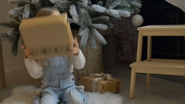 Söt blond flicka har fått en presentförpackning insvept i hantverk papper sitter nära julgran, lycklig barndom och jul firande koncept — Stockvideo