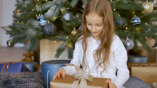 Schattig blond meisje heeft ontvangen een geschenkdoos verpakt in ambachtelijk papier zitten in de buurt van kerstboom, gelukkige kindertijd en Kerstmis viering concepten — Stockvideo