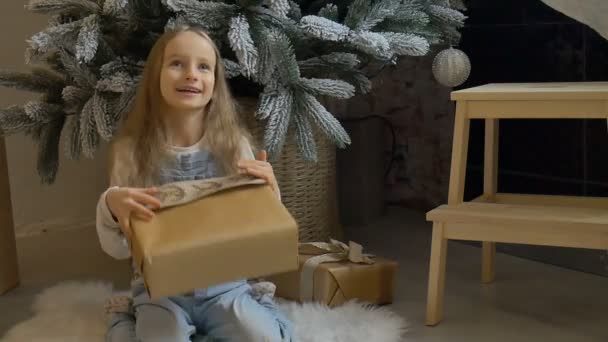 Linda chica rubia ha recibido una caja de regalo envuelta en papel artesanal sentado cerca del árbol de Navidad, la infancia feliz y los conceptos de celebración de Navidad — Vídeo de stock