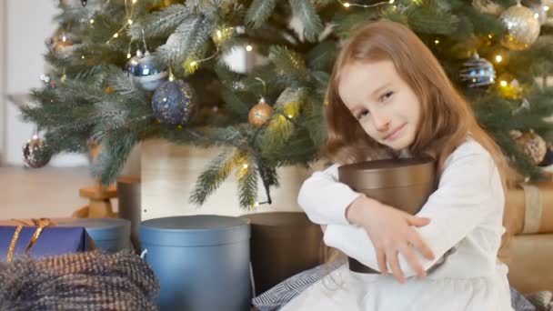 Gülümseyen sarışın kız, aldığı altın hediye kutusuna sarılıyor. Noel ağacı, mutlu çocukluk ve xmas kutlaması kavramlarının yanında oturuyor. — Stok video