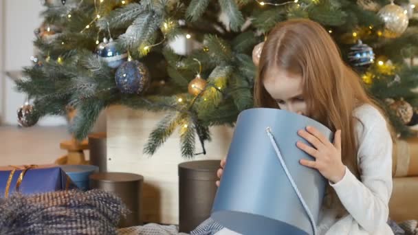 Menina loira bonito está abrindo caixa de presente azul escuro que ela recebeu sentado perto de árvore de natal, infância feliz e conceitos de celebração de xmas — Vídeo de Stock