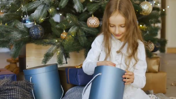 Şirin sarışın kız aldığı koyu mavi hediye kutusunu açıyor. Noel ağacının yanında oturuyor. Mutlu çocukluk ve xmas kutlaması kavramları. — Stok video