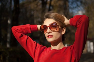 Güneş gözlüğü takan ve kırmızı kazak giyen güzel bir kadın açık havada güneşli bir havada sonbahar parkında poz veriyor.
