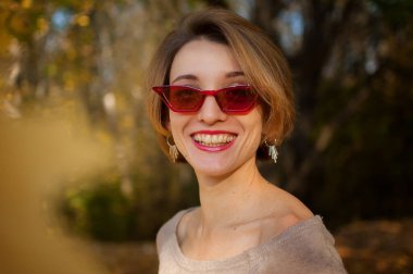 Genç ve güzel bir kız, kısa saçlı ve kırmızı gözlüklü sarı yapraklı ağaçlara karşı poz veriyor sonbahar parkında vakit geçiriyor.