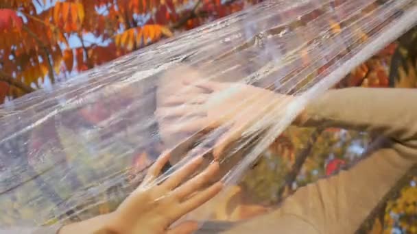 Молодая женщина с короткими светлыми волосами смотрит через пластиковый пакет, полиэтиленовая пленка стоит на открытом воздухе durind солнечный осенний день рядом с деревом с желтыми и красными листьями . — стоковое видео