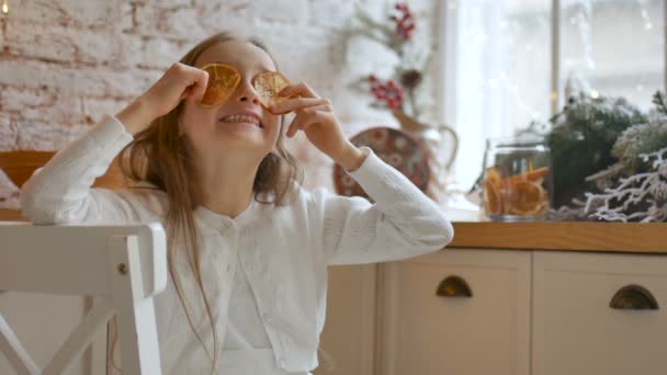 Lachend blond meisje grimmig en bedekt haar ogen met gedroogde sinaasappelschijfjes thuis met loft stijl en kerstversiering op achtergrond, vrolijk kerstconcept — Stockvideo