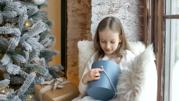 Menina loira bonito está abrindo caixa de presente azul escuro que ela recebeu sentado perto de árvore de natal, infância feliz e conceitos de celebração de xmas — Vídeo de Stock