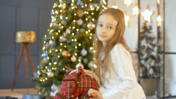 Nettes blondes Mädchen reitet auf einem Spielzeug Schaukelpferd vor dem Weihnachtsbaum und der Wand mit gelben Sternen, Neujahr 2020, Konzept für Weihnachten — Stockvideo