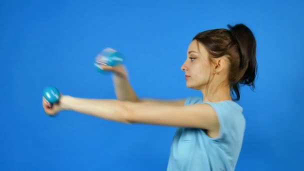 Pretty Young Sports Woman with Dumbbell in Hand Making Her Exercise at Blue Studio Background (en inglés). Chica bonita con cola de caballo en Fitwear. Concepto de culturismo . — Vídeos de Stock