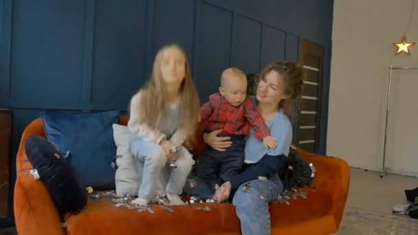 Família feliz celebra com ouro e prata confete em casa sentado no sofá laranja no fundo da parede azul escuro, sorrindo filha pulando e jogando pequenos pedaços de confete — Vídeo de Stock