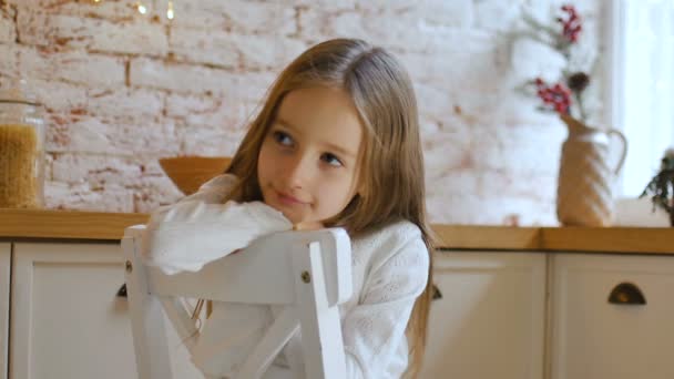 Liten ledsen flicka med blå ögon och långt blont hår bär vit klänning och tröja sitter på en stol i en loft stil rum — Stockvideo