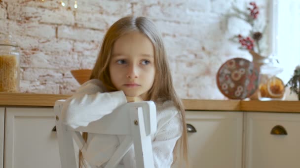 Niña triste con ojos azules y pelo largo y rubio con vestido blanco y suéter se sienta en una silla en una habitación estilo loft — Vídeos de Stock