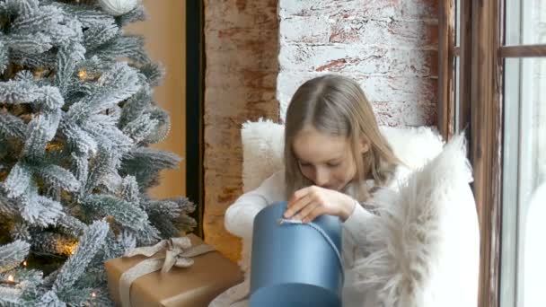 Симпатична блондинка відкриває темно-синю подарункову коробку, яку вона врятувала, сидячи біля ялинки, щасливого дитинства та різдвяних святкувань — стокове відео