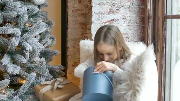 Linda chica rubia está abriendo la caja de regalo azul oscuro que recibió sentado cerca del árbol de Navidad, la infancia feliz y los conceptos de celebración de Navidad — Vídeo de stock