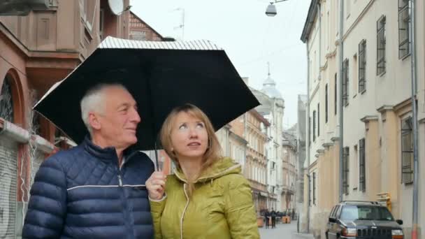 Frau mittleren Alters und ihr älterer Mann verbringen die Zeit zusammen im Freien unter ihrem Sonnenschirm auf gepflasterter Straße. Paar mit Altersunterschied. — Stockvideo