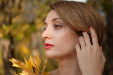 Sarışın, kısa saçlı, güzel, kırmızı-altın bir sonbahar parkında elinde bir buket yaprak tutan çekici genç bir kadın.